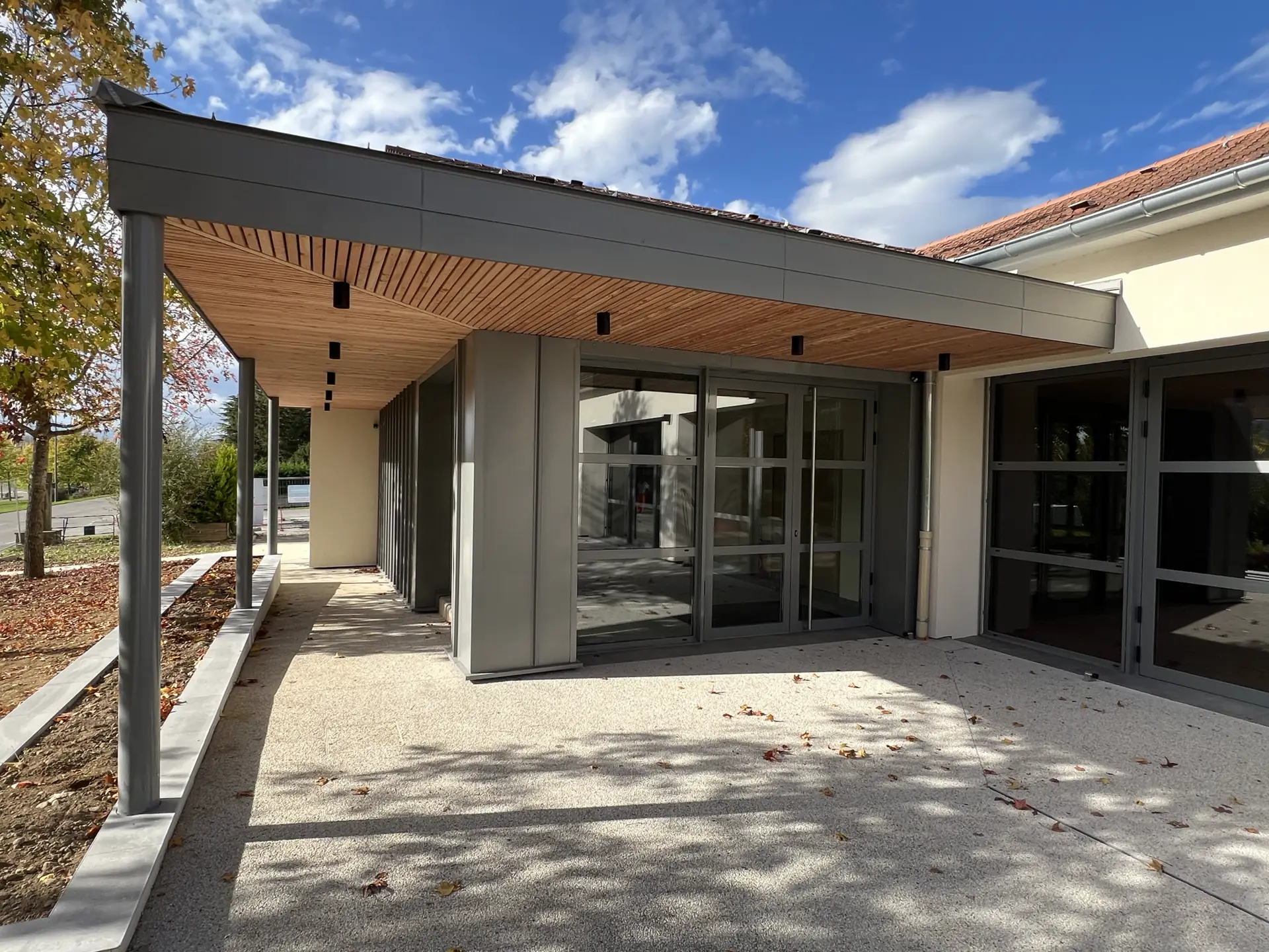 Entrée vitrée d'une salle communale sous un préau à sous-face en bois à Vernioz